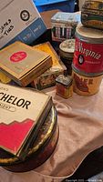 Photo showing a variety of vintage cigar boxes and tobacco tins on a beige surface, including Old Virginia and Admiral Schelor items.