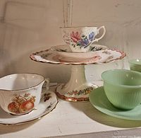 Photo showing collection of milk glass cups and saucers, fine china teacups and saucers, and ceramic floral cake plates on pedestal stands.