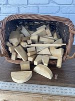 Basket filled with oversized wooden building blocks including various geometric shapes like arch, wedge, cylinder, rectangular and small peg pieces placed against a brick background with a ruler showing scale.