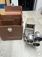 Front view of Bell & Howell 3 lens 8mm camera next to brown leather carrying case on wood floor.
