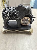 Front view of black elephant figurine holding clock with Roman numeral face and gold bezel, standing on a decorative gold and black base.