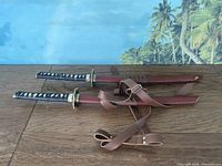 Pair of cosplay samurai swords laid side by side showing red scabbards and brown leather carry straps.