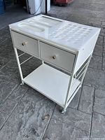 Front and side view of white craft vanity table stand showing molded top compartments, two drawers, shelf, and wheels