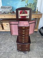 Full view of dark wood, tall and narrow jewelry cabinet with fliptop lid and mirror. Sides open with hooks for hanging necklaces. Eight drawers shown with brass handles.