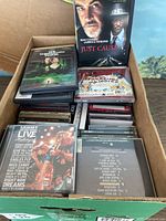 Full view of the box containing multiple DVDs, CDs, and cassette tapes arranged loosely with some CDs visible with titles and DJs' names, and small glimpses of cassette tape cases