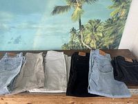 Six pairs of pants arranged in a row showing color variations: light blue, grey, off-white, black, light blue, and brown corduroy shorts.