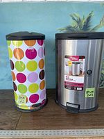 Pair of foot kick garbage cans shown side by side on a wooden surface with a backdrop. One has colorful polka dots and the other is stainless steel with a lid.