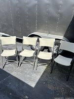 Four beige folding chairs with black metal frames outside on pavement by a trailer tire.