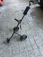 Black golf push cart standing on a textured concrete floor, showing angled front view with three wheels and adjustable handle.