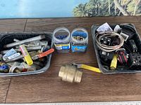 Top-down image showing three containers with assorted plumbing fittings including metal valves, pipe pieces, small plumbing connectors, and plastic containers with small parts such as screws or pins.