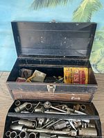 Black metal toolbox open showing interior with various hand tools and papers inside.