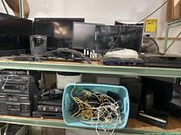 Wide view of two shelves holding various electronics including TVs, monitors, a blue bin filled with assorted cables, stereo components, and VCRs