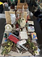 Zoomed out view of a large 8x4 table filled with miscellaneous household items and decor including vases, a fan, boxed goods, and decorative greenery.