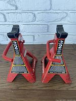 Pair of red Husky 2 ton jack stands with handles and safety labels on a wooden surface against a white brick wall.