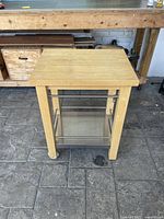 Front view of wooden butcher block kitchen island with metal bars and wheels on stone floor showing used condition.