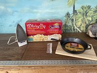 Photo showing white iron, Whirley Pop popcorn popper box, Lodge Logic cast-iron skillet on wooden cutting board, and small packet.