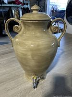 Front view of large urn-shaped water jug with lid and metal spigot at the base. Two curled handles visible.