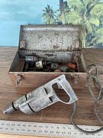 Milwaukee heavy duty drill and open rusty metal carry case showing multiple drill bits inside, drill cord coiled beside it, measuring tape in background for scale.