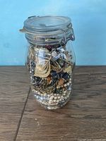Front view of full glass jar containing mixed jewelry pieces, showing layered contents including beads, metal chains, and pins.