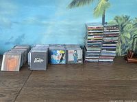 Wide view of multiple stacks of music CDs in jewel cases on a wooden table in front of a tropical wall mural.