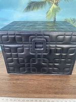 Front view of black padded storage chest with stitch pattern and a decorative square clasp, placed on wooden surface with a ruler in front measuring approximately 20 inches in width.