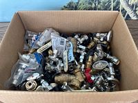 Box filled with various plumbing shut off valves and connectors, including metal valves with handles in brass and chrome finish and plastic fittings.