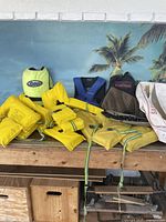 Full lot showing various yellow lifejackets, a lime green Sterns lifejacket, a brown mesh fishing vest, and a white bag, arranged on a wooden surface with a tropical backdrop.