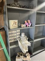 Photo of shelf containing white ceramic dishes stacked in pile, two baby dolls dressed in pink, small white boxes, and framed decoration box.