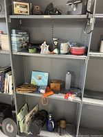 Metal storage shelves displaying a variety of household items including books, ceramic decorations, mugs, a water bottle, gloves, wooden boxes, and small containers.