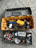 Opened yellow toolbox with black lid showing inside compartments filled with assorted tools including a yellow drill attachment, pliers, and various plumbing fittings and hardware.