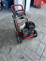 Front angle view showing Troy-Bilt branding, red frame, black engine, and attached hose with spray gun on paved surface.