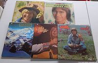 Five John Denver LP vinyl records with their covers, arranged in a 3-2 layout. Covers show images including a smiling John Denver with a hat, a close-up portrait, snow-covered landscape, John Denver smiling in a close shot with a guitar, and John Denver sitting on grass.