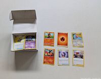 Open box of assorted Pokemon cards with several cards displayed outside the box, showing a mix of character and energy cards.