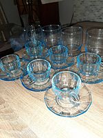 View showing six blue depression glass espresso cups each paired with matching saucers arranged on a wooden surface along with four taller blue depression glass water/highball glasses behind them.