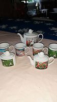 Group photo showing entire 7-piece ceramic tea service set with teapot, sugar bowl, creamer, and 4 mugs, each decorated with colorful Deco style whimsical illustrations. White ceramic base with glossy finish.