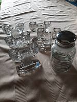 Five clear glass double candleholders and one clear glass jar with silvered stopper displayed on a table to show full lot contents