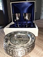 Photo of stacked 13 silver plated plates with embossed grapevine designs and 2 boxed silver plated goblets on blue satin lining.