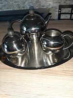 Photo from a slightly elevated angle showing the full tea service set including the teapot with lid, sugar bowl with lid, creamer, spoon on an oval tray all made of chrome.