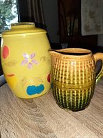 Photo showing a yellow vintage cookie jar with painted flower and fruit designs next to a brown and green ceramic milk jug with textured ribbed pattern and handle on a wooden surface.