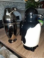 Photo showing both the penguin thermos and chrome cocktail shaker side by side on a wooden surface under natural lighting.