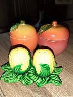 Close-up photo showing one set of vintage ceramic salt and pepper shakers shaped like yellow corn on green leaves and a second set of matching ceramic marmalade pots shaped like peaches or nectarines in orange and pink hues with leaf-shaped lids.