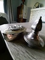 Photo showing vintage silver plate trinket box with floral embossed lid and a silver plate candle snuffer with bulbous handle.