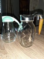 Two vintage syrup jugs placed side by side on a wooden surface. Left jug has mint green plastic lid and handle; right jug has metal lid and brown plastic handle.