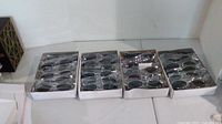 Four boxes visible, each containing 12 oval sunglasses, with plastic wrap over them. Sunglasses have dark lenses and metallic frames.