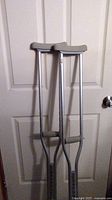 Two aluminum adjustable crutches leaning against a white door, showing height-adjustment holes and padded grips.