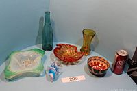 Photo showing orange bowl, green and white dish, blue glass bottle, small blue and white fish figurine, amber goblet, and small brown and orange bowl together on table.