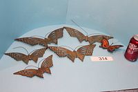 Five brass butterflies and one small painted butterfly placed on a blue surface next to a soda can for size reference.