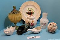 Wide view showing all seven assorted decorative items including butterfly plate, two floral jars, two vases, lidded green glass jar, feeding cup and black dish.