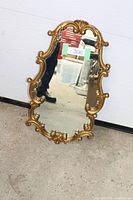 Front view of the ornate gold framed wall mirror placed on the floor against a white wall, showing detailed scrolling and floral design of frame.