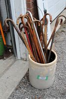 Photo showing a large ceramic crock containing several used wooden canes with curved handles, set outside near a garage door.